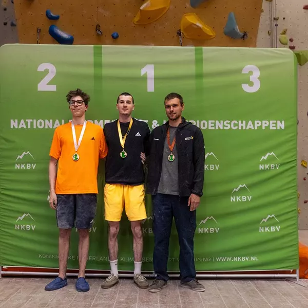 Drie mannen staan op een podium met medailles om, voor een groene achtergrond waarop ‘NKBV’ en ‘Nationale Kampioenschappen’ staat geschreven. Achter hen zijn de podiumnummers 2, 1 en 3 te zien.